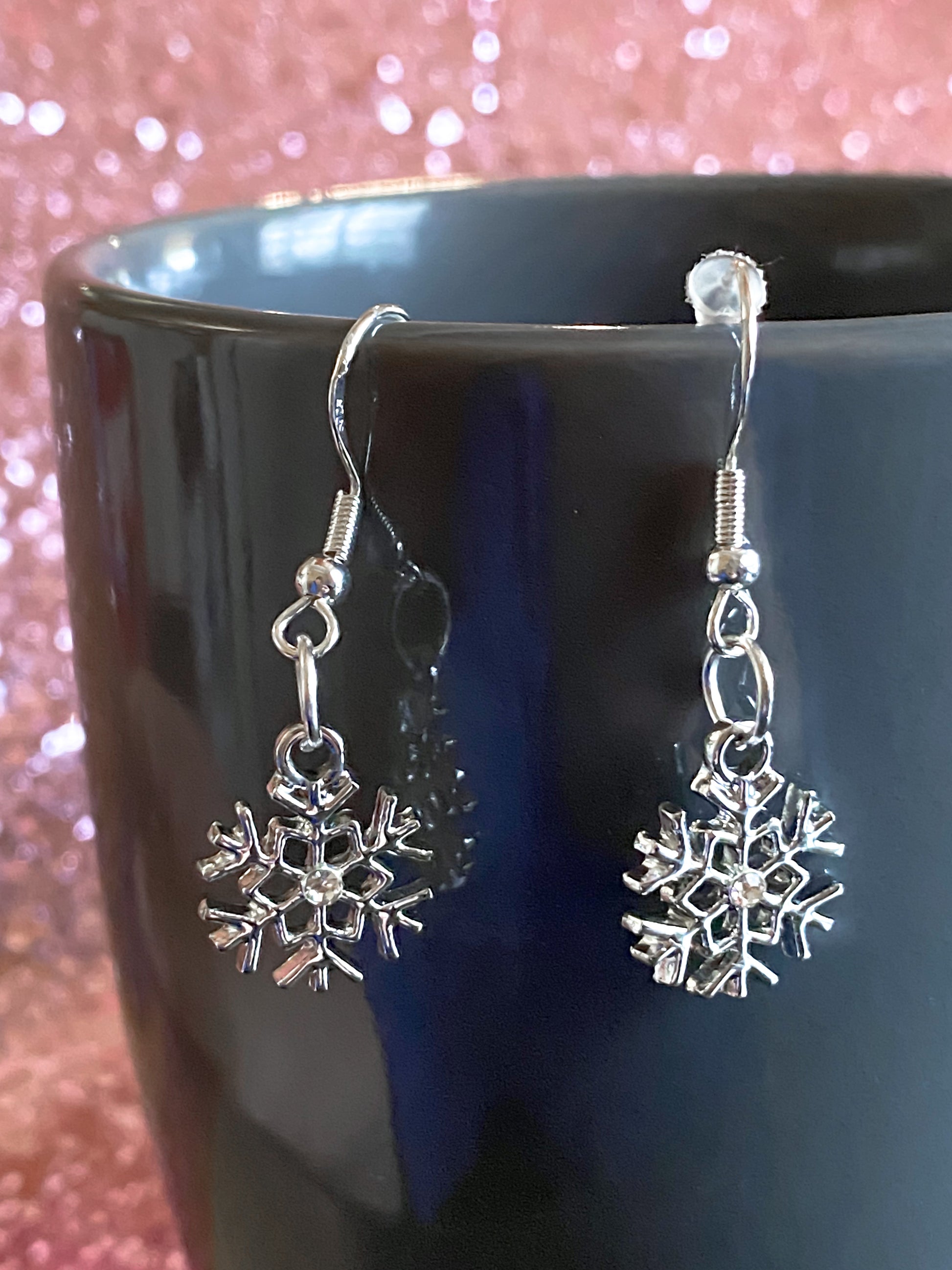Silver snowflake dangle earrings with rhinestone in the middle, shown handing from a gray coffee mug with pink glitter background. Hooks show 925 on them, signifying they are hypoallergenic