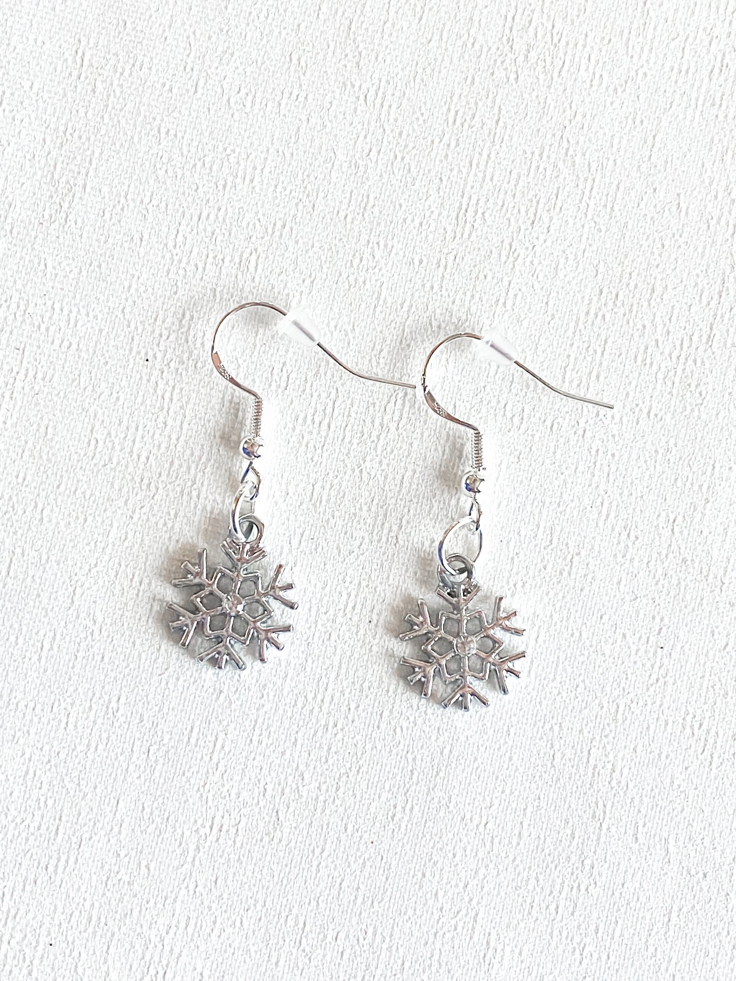 Silver dangle snowflake earring with crystal rhinestone in the middle of the earring piece. Set against white textured background