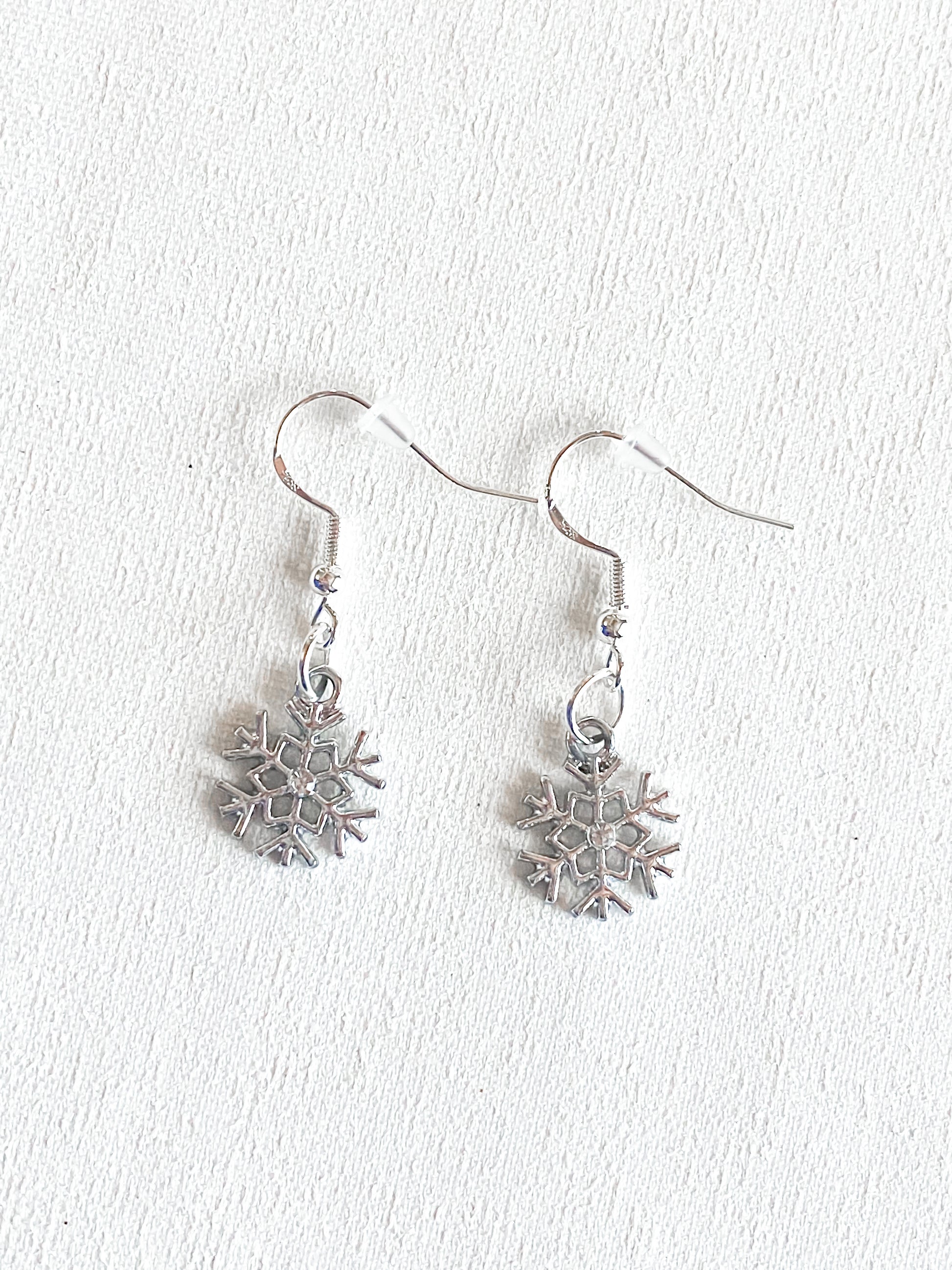 Silver dangle snowflake earring with crystal rhinestone in the middle of the earring piece. Set against white textured background