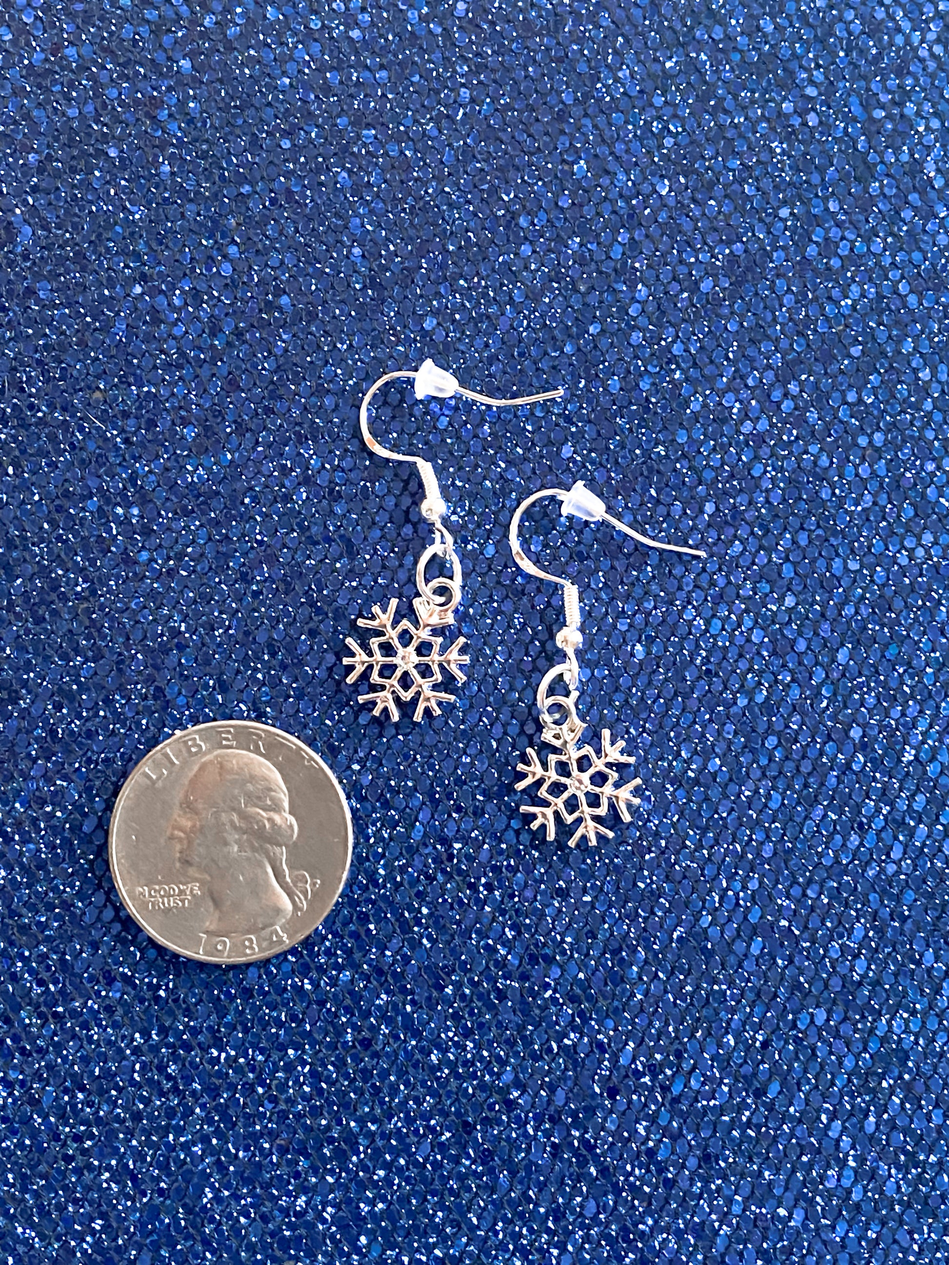 Crystal Rhinestone silver snowflake dangle earrings against blue textured shimmer and glitter background. Quarter shown to compare size of earrings. these earrings about half the size of the quarter.