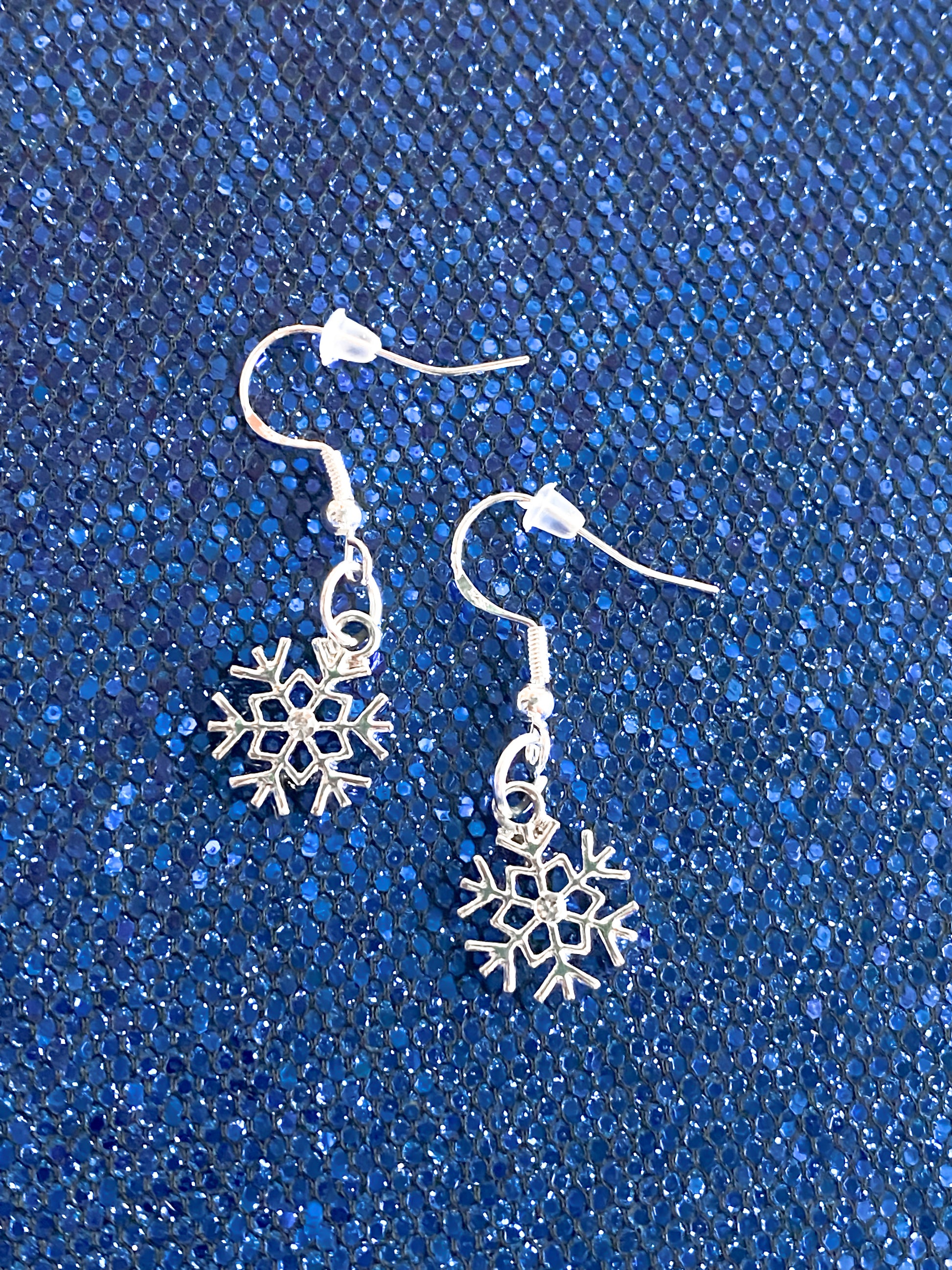 Pretty silver snowflake dangle earrings set against blue textured shimmer background. Earrings have a crystal in the middle, hooks are silver, includes a clear rubber back piece