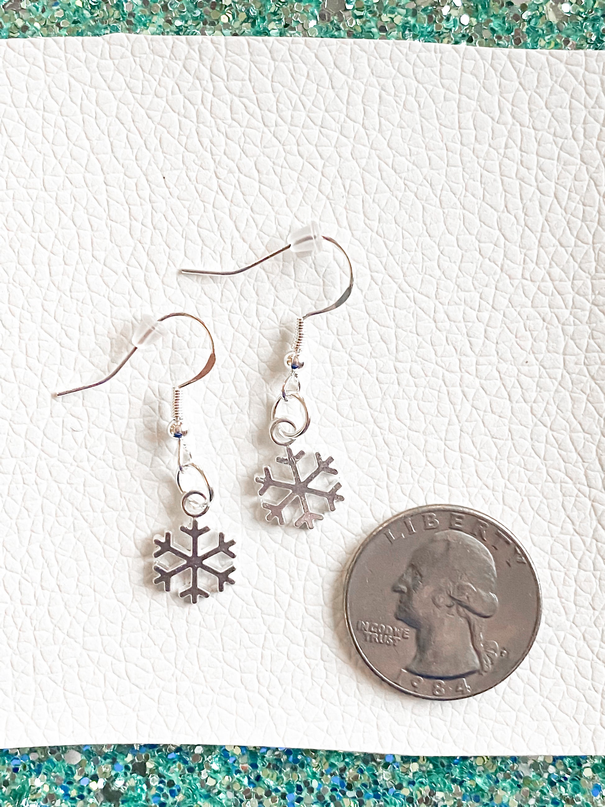 Small snowflake earrings set against white leather and teal glitter. Quarter on bottom right hand side to show sizing - both of these earrings could fit within the quarter.