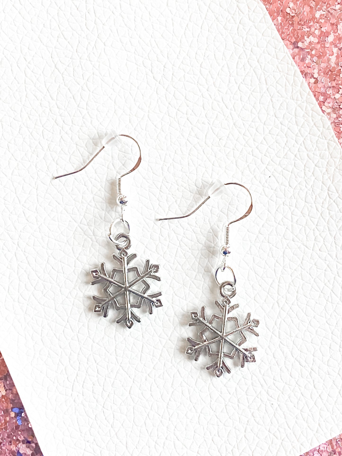 Silver dangle snowflake earrings against white leather with some pink glitter. Silver snowflakes have silver hooks and rubber safety back