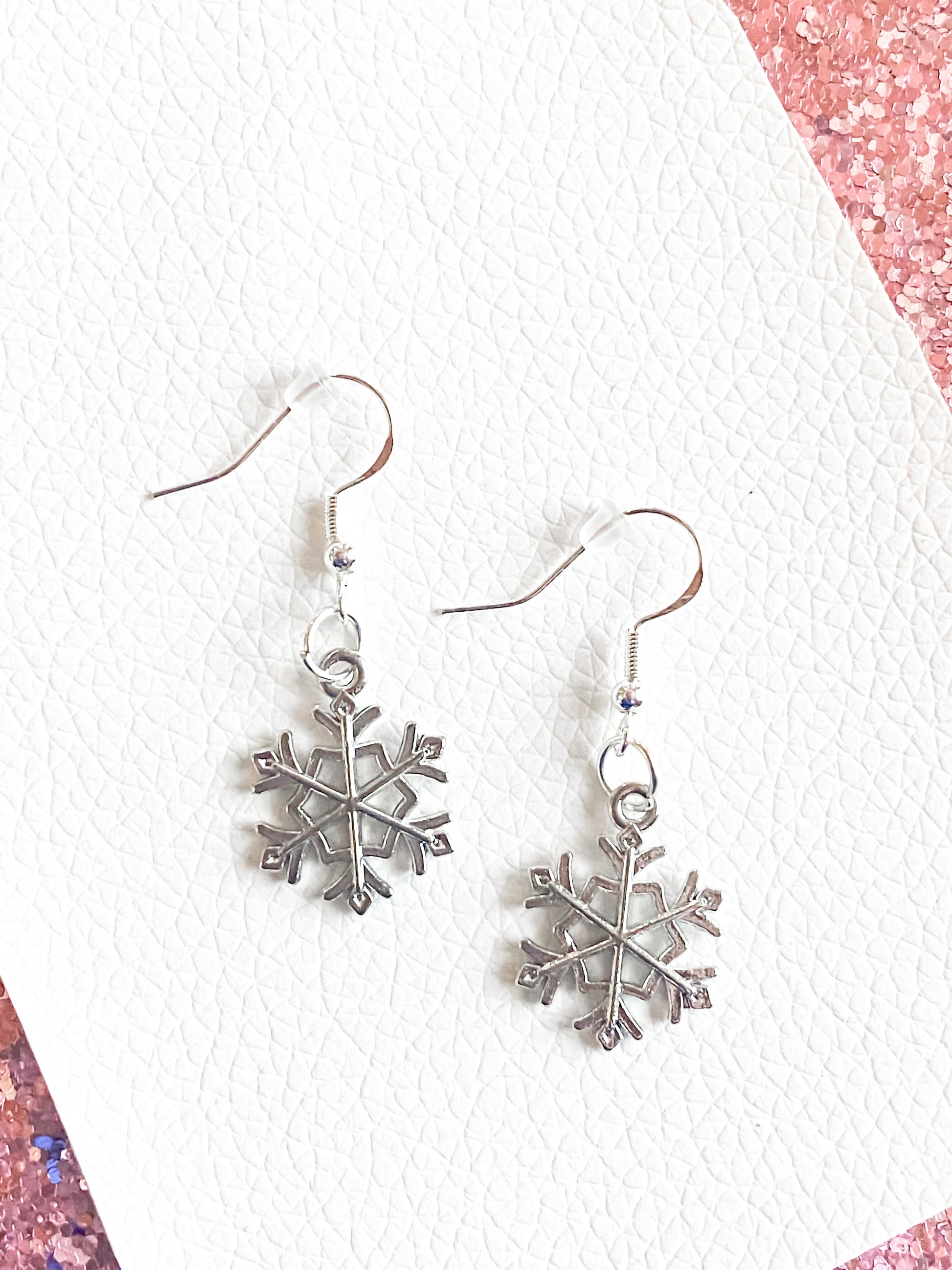 Silver dangle snowflake earrings against white leather with some pink glitter. Silver snowflakes have silver hooks and rubber safety back