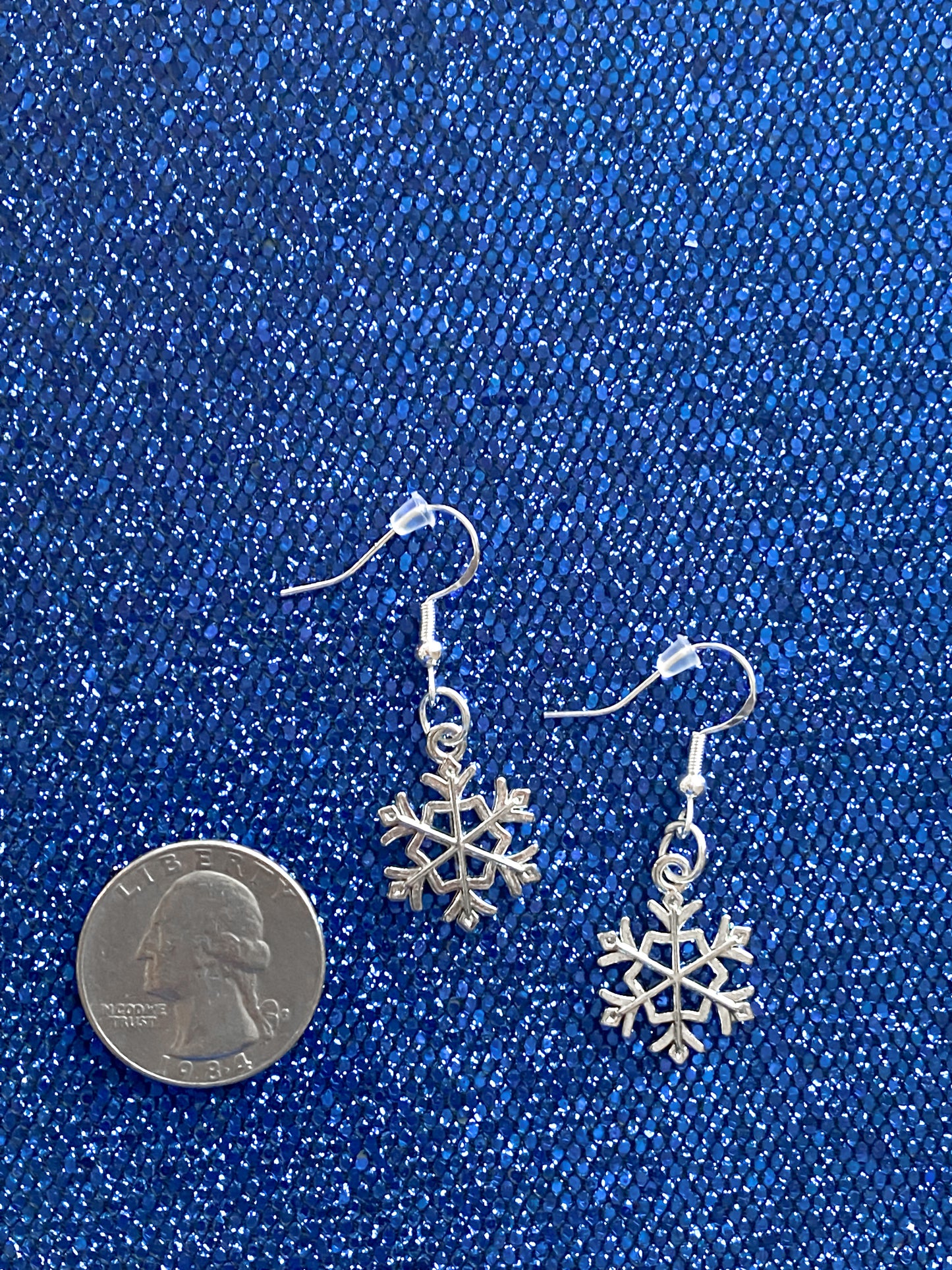 Silver snowflake dangle earrings against blue glitter background with a quarter nearby to show relative size of earrings. A single snowflake earring is smaller than the size of a quarter