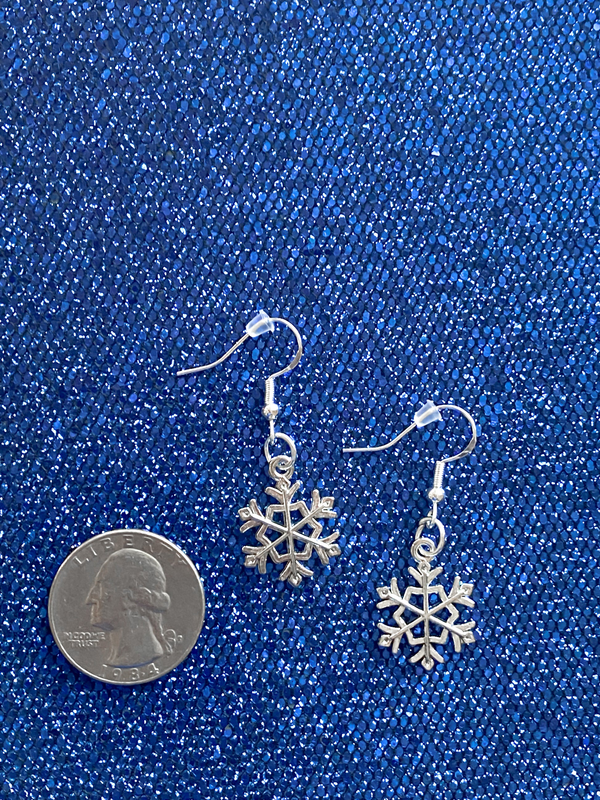 Silver snowflake dangle earrings against blue glitter background with a quarter nearby to show relative size of earrings. A single snowflake earring is smaller than the size of a quarter